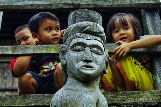 Kids peeping out behind a Dayak Hampatong wooden pole sculpture